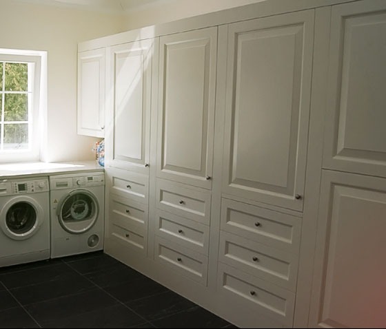 Białe szafy z szufladami w zabudowie, dopasowane do pralni z dwoma pralkami obok okna, czarna podłoga.
