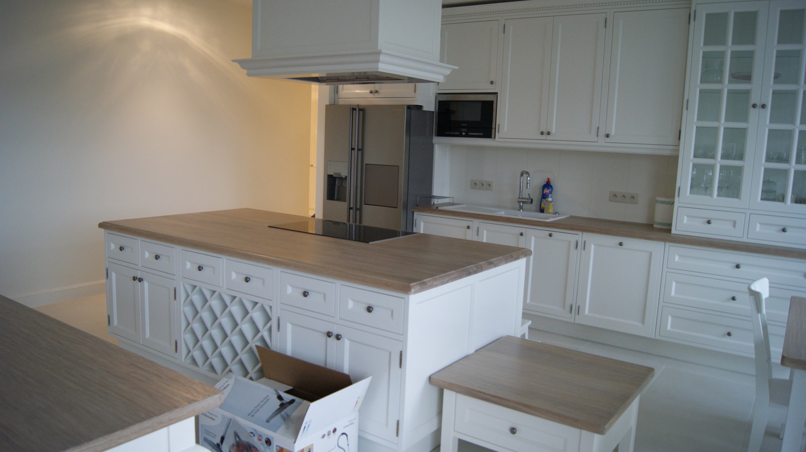 Jasna kuchnia z białymi szafkami i drewnianym blatem, wyspa kuchenna z kratownicą na wino i pudełkiem po sprzęcie AGD na podłodze.
