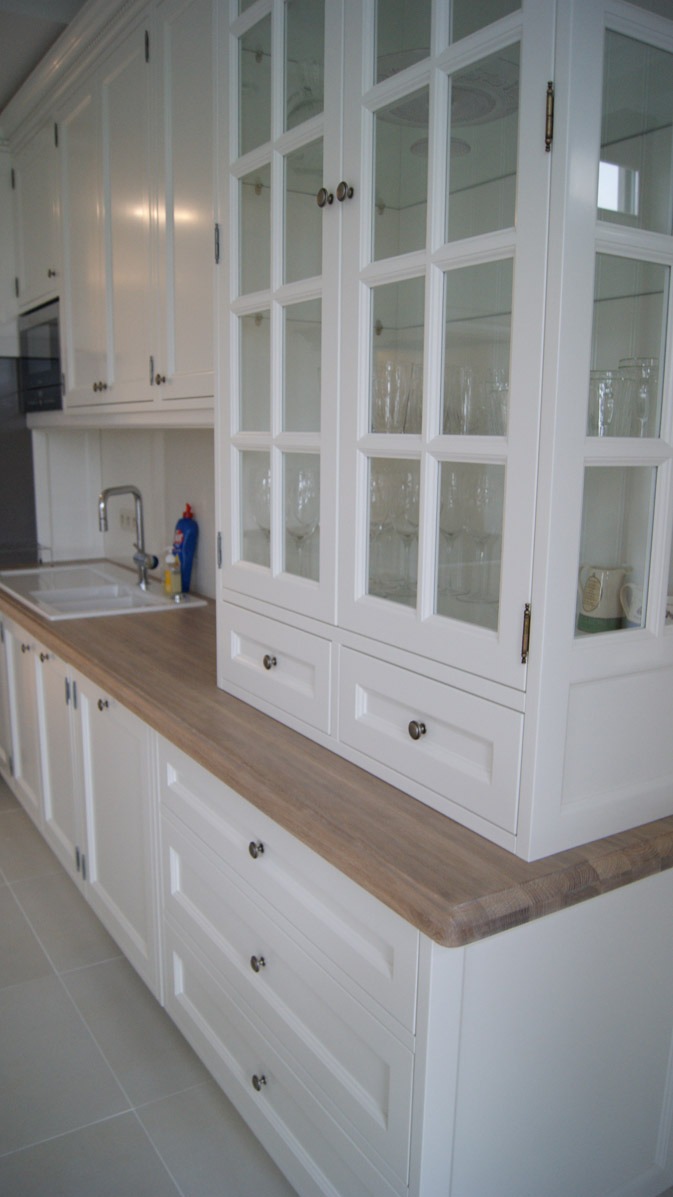 Biała zabudowa kuchenna z drewnianym blatem, przeszklona witryna z dekoracyjnymi uchwytami i szklankami w środku, widoczny zlew i dozownik płynu.