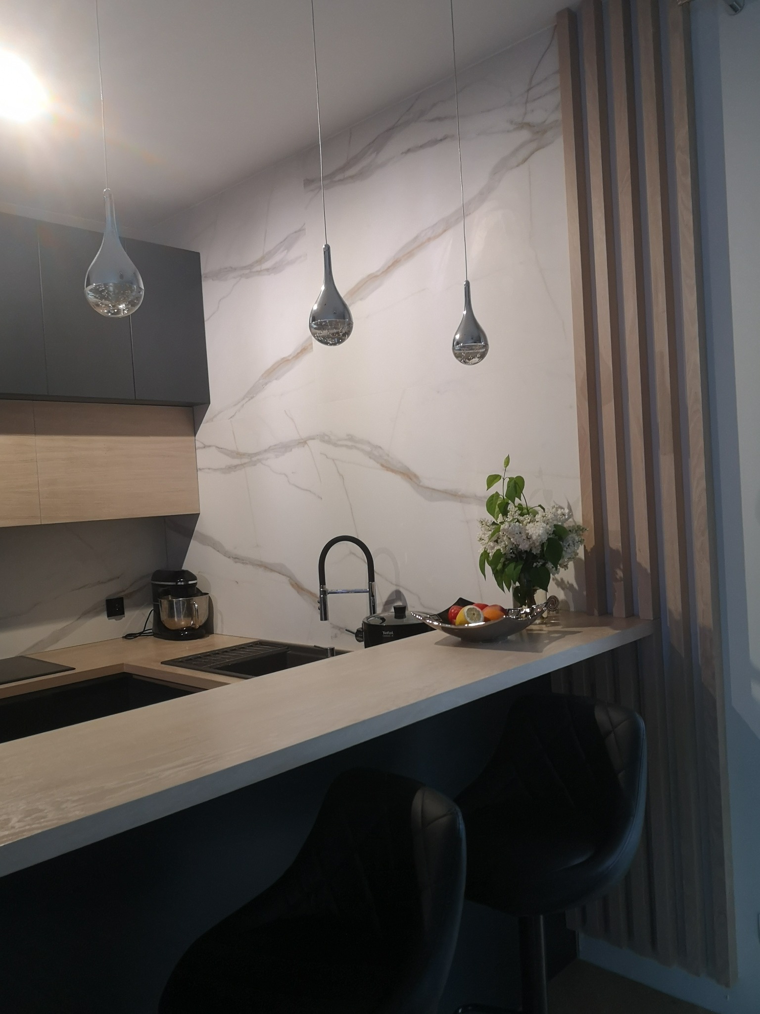 Nowoczesna kuchnia z marmurowym blatem, czarnymi krzesłami barowymi, minimalistycznymi lampami wiszącymi i dekoracyjną ścianką z drewnianych listew.