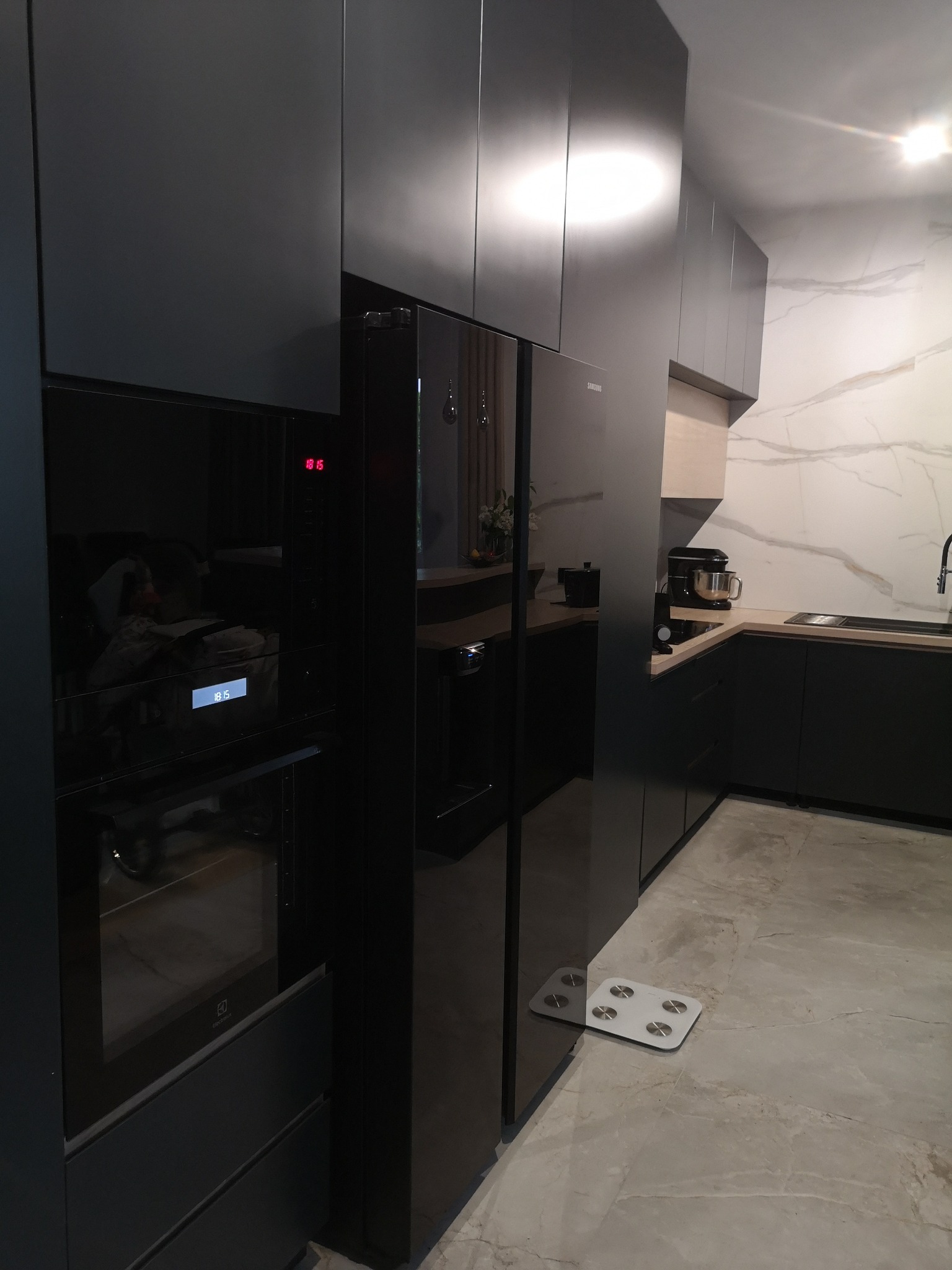 Nowoczesna, czarna zabudowa kuchenna z piekarnikiem i lodówką side-by-side, uzupełniona marmurową ścianą i wagą łazienkową na podłodze.