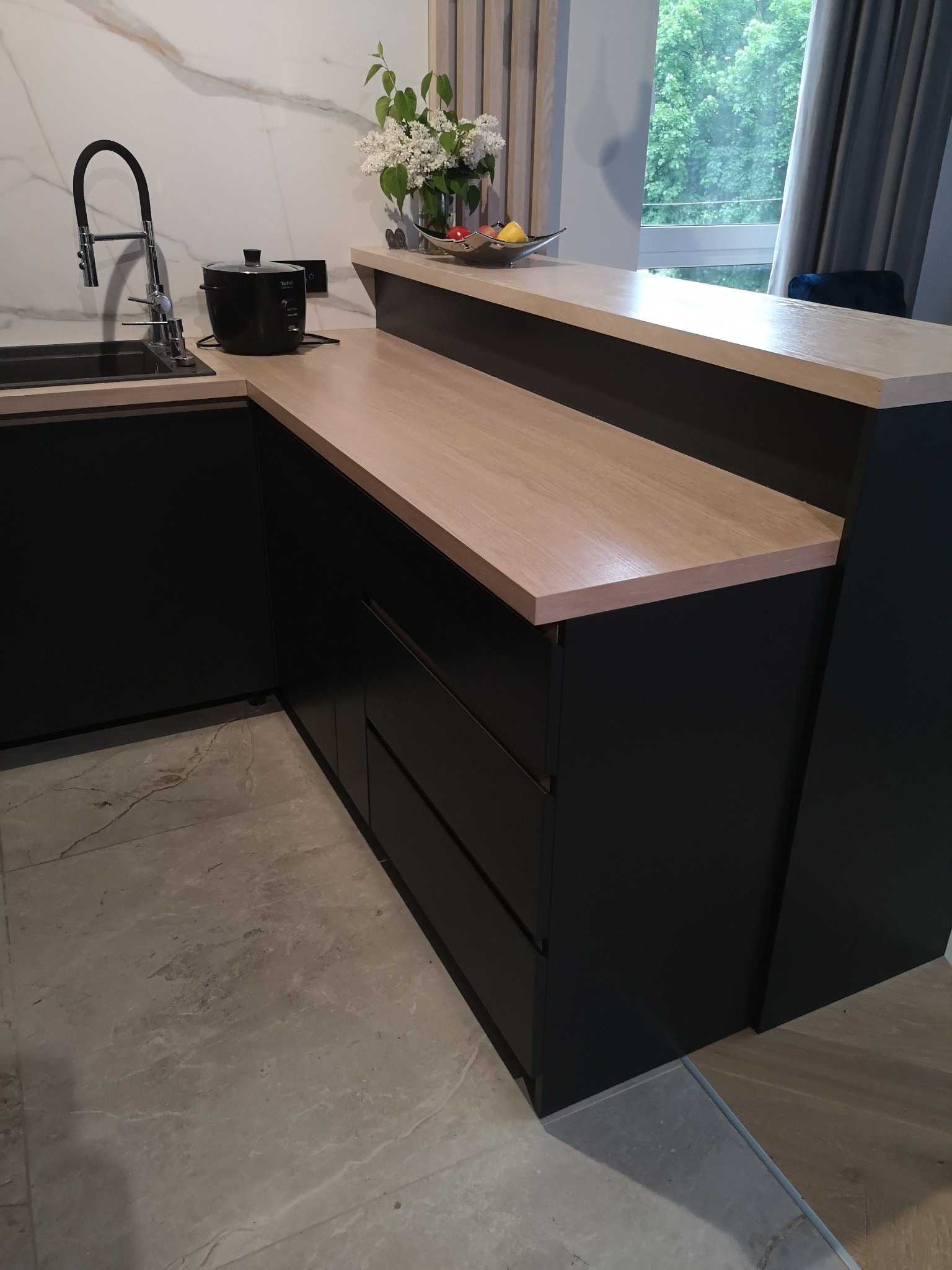 Nowoczesna kuchnia z czarnymi szafkami i blatem w kolorze jasnego drewna, z widocznym zlewem, baterią i sprzętem AGD na blacie, marmurowa ściana w tle i kwiaty w wazonie.