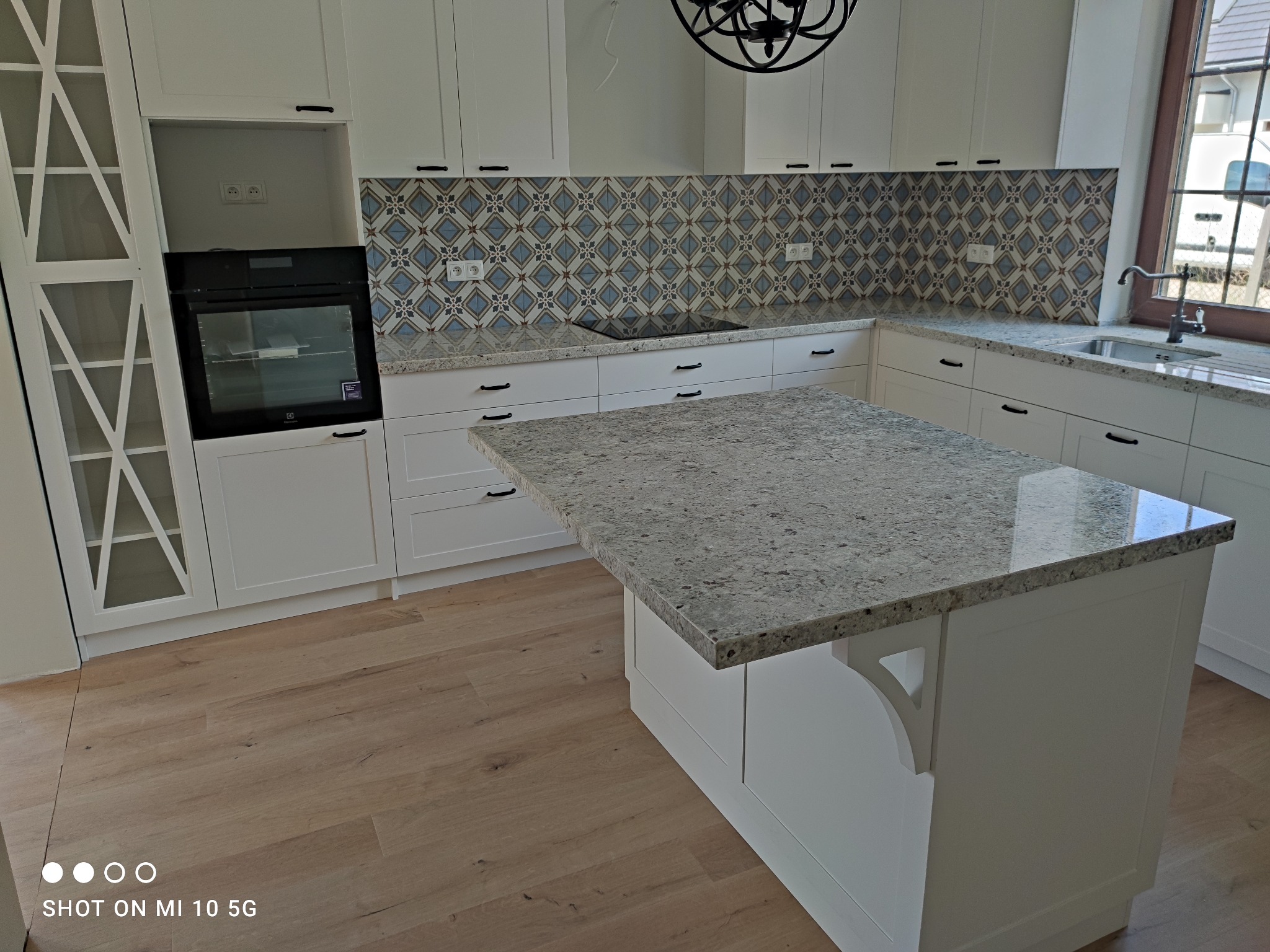 Jasna kuchnia z wyspą i wzorzystą mozaiką nad blatem, widoczny piekarnik w zabudowie i drewniana podłoga.