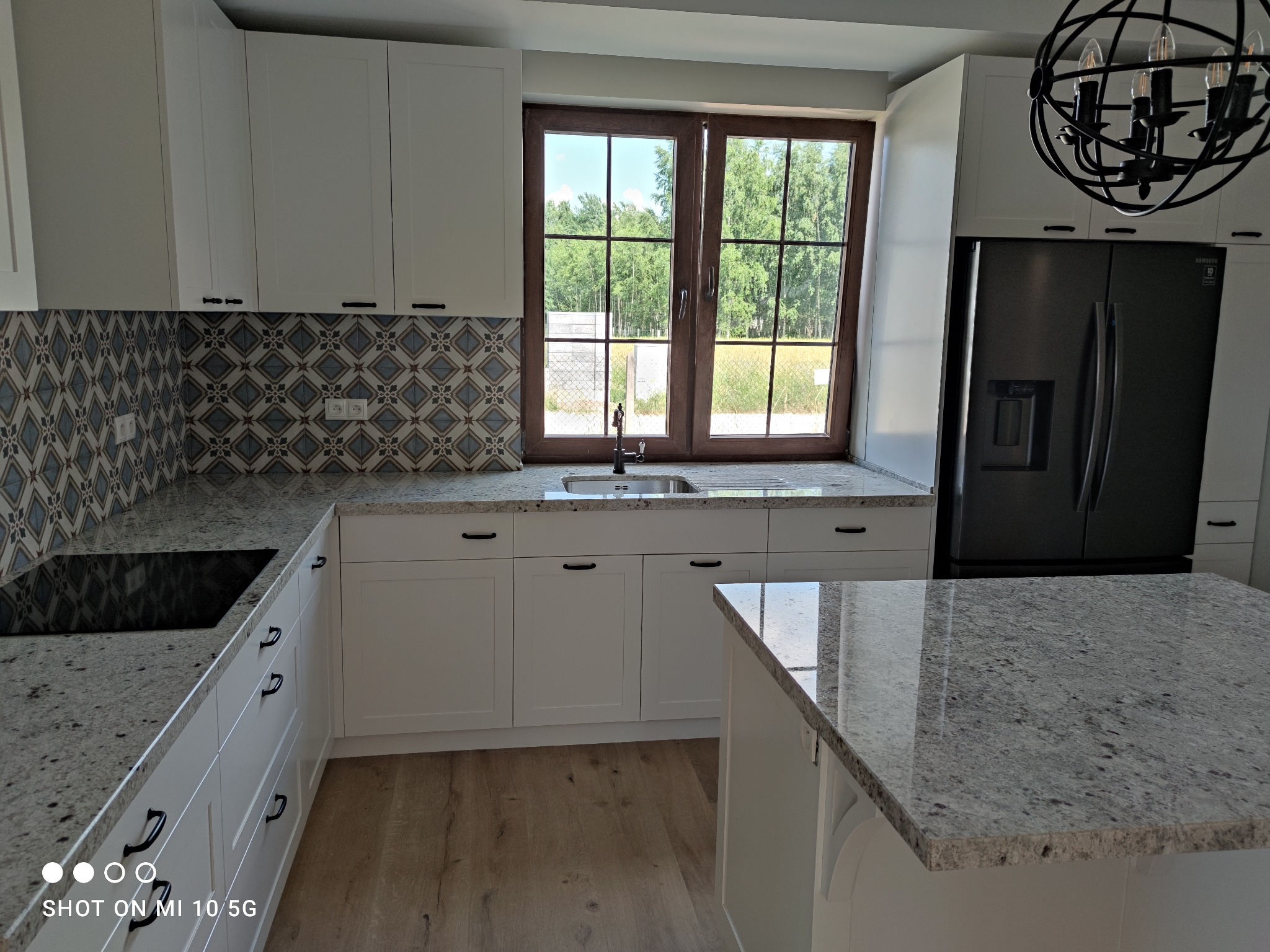 Nowoczesna kuchnia z białymi szafkami, blatem z jasnego granitu i dekoracyjną mozaiką nad blatem. Widoczne okno z widokiem na zieleń, czarna lodówka i lampa w stylu industrialnym.