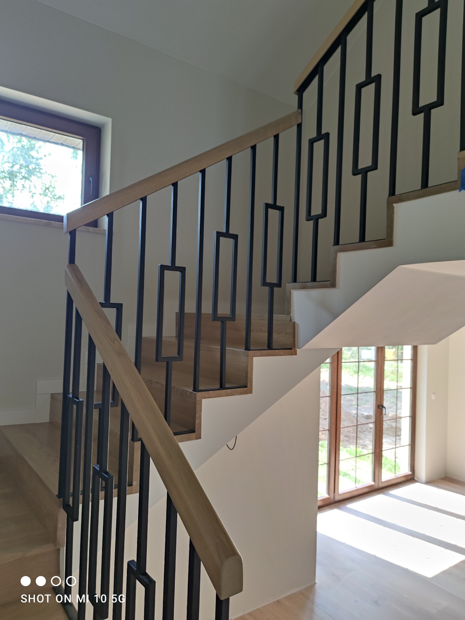 Nowoczesne schody z drewnianymi stopniami i balustradą z czarnymi, prostokątnymi elementami, oświetlone naturalnym światłem wpadającym przez okno.