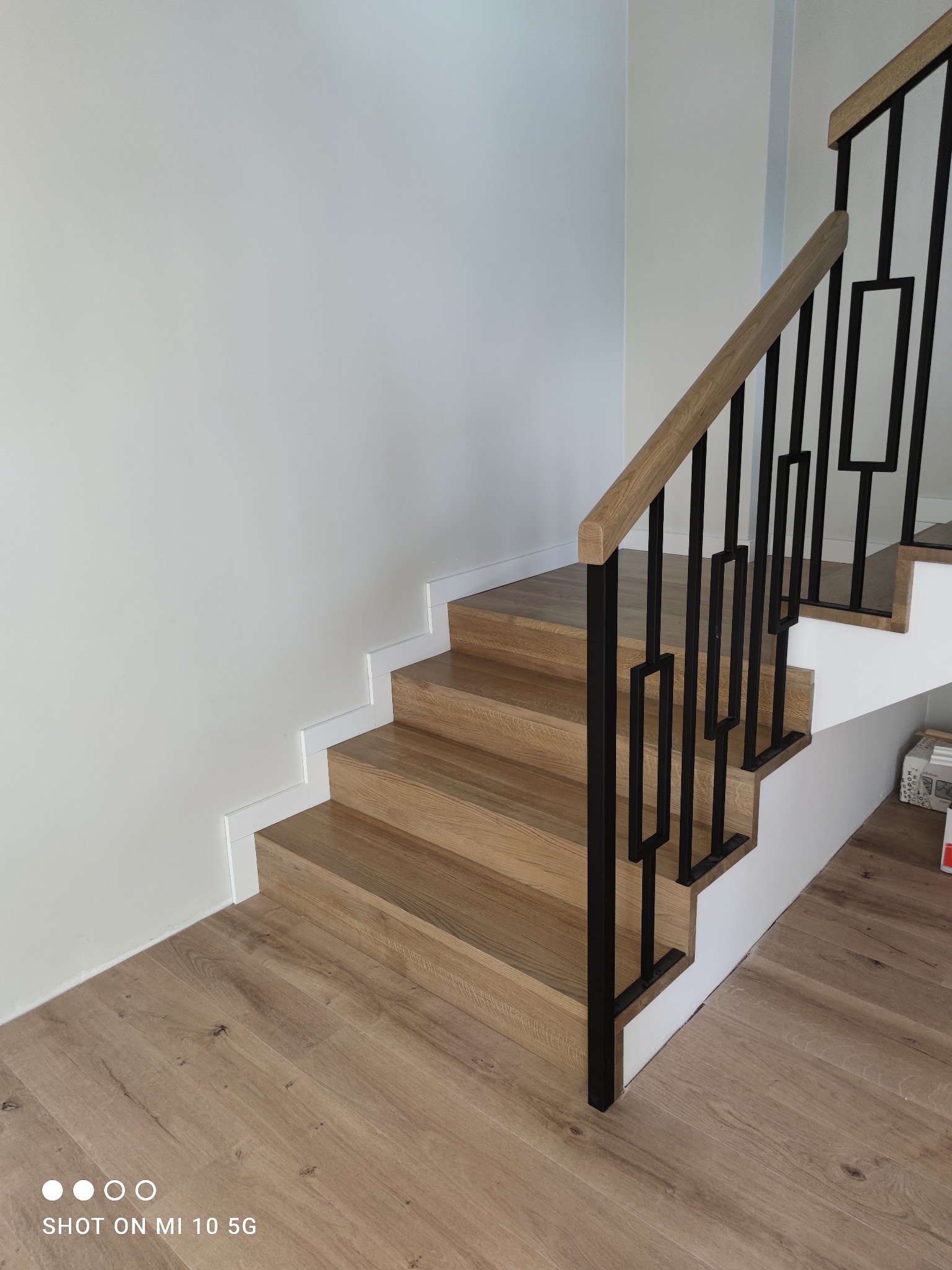 Jasne, drewniane schody z czarną, geometryczną balustradą i białymi listwami przypodłogowymi, prowadzące w górę, widoczne z dołu.