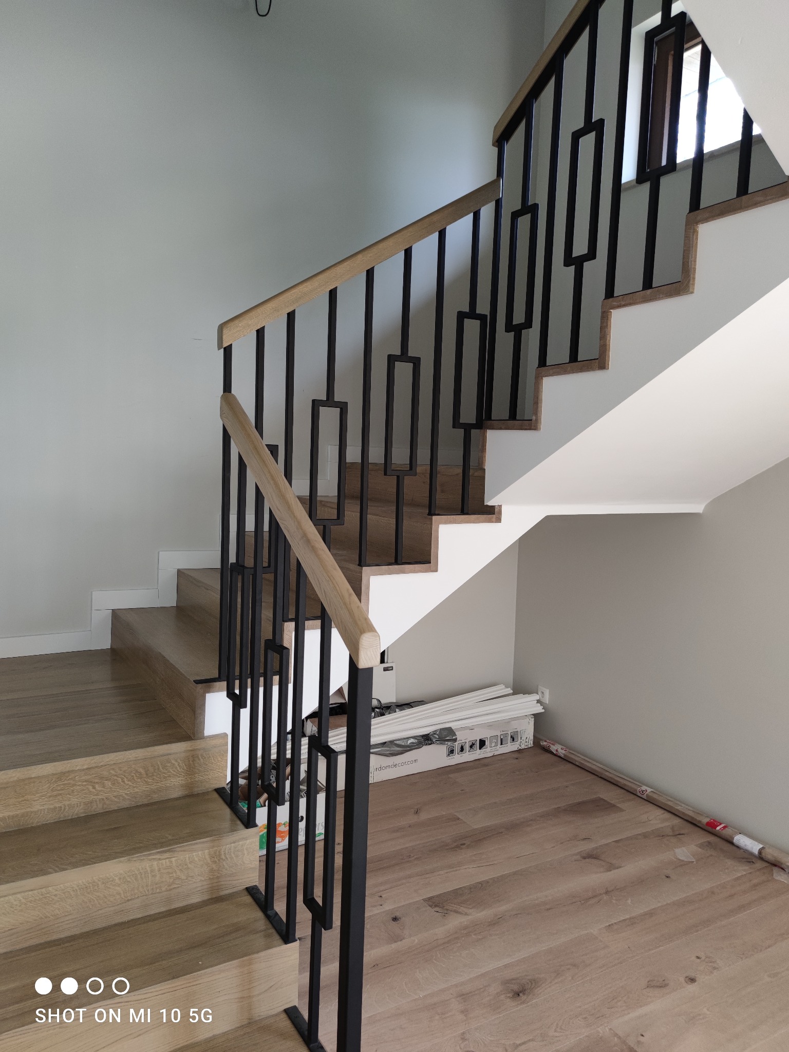 Nowoczesne schody z drewnianymi stopniami i czarną, geometryczną balustradą, częściowo zakryte opakowaniami materiałów budowlanych, na tle szarej ściany.