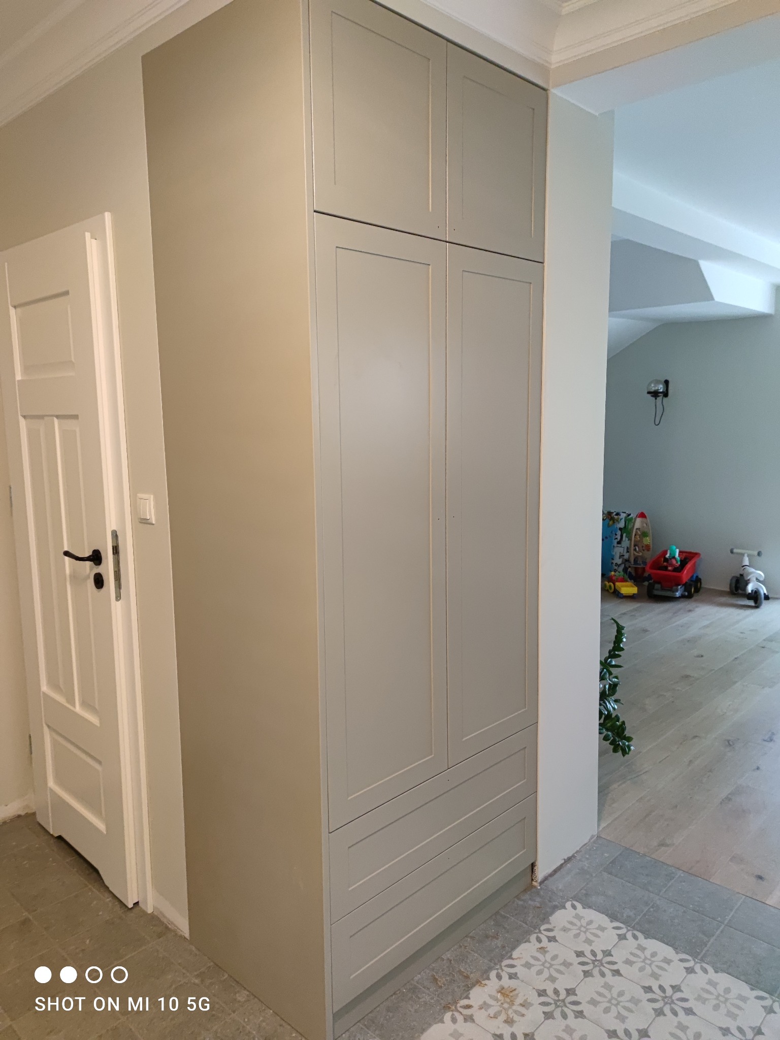 Wysoka, szara szafa wnękowa z frezowanymi frontami, dopasowana kolorystycznie do ściany, z widocznymi trzema szufladami w dolnej części i białymi drzwiami po lewej stronie, w tle pokój dziecięcy...