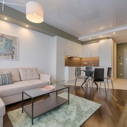 Jasne wnętrze: beżowa sofa z poduszkami, szklany stolik kawowy na tle kuchni z białymi szafkami i drewnianym blatem. Całość dopełnia zielony dywan i obraz na ścianie.