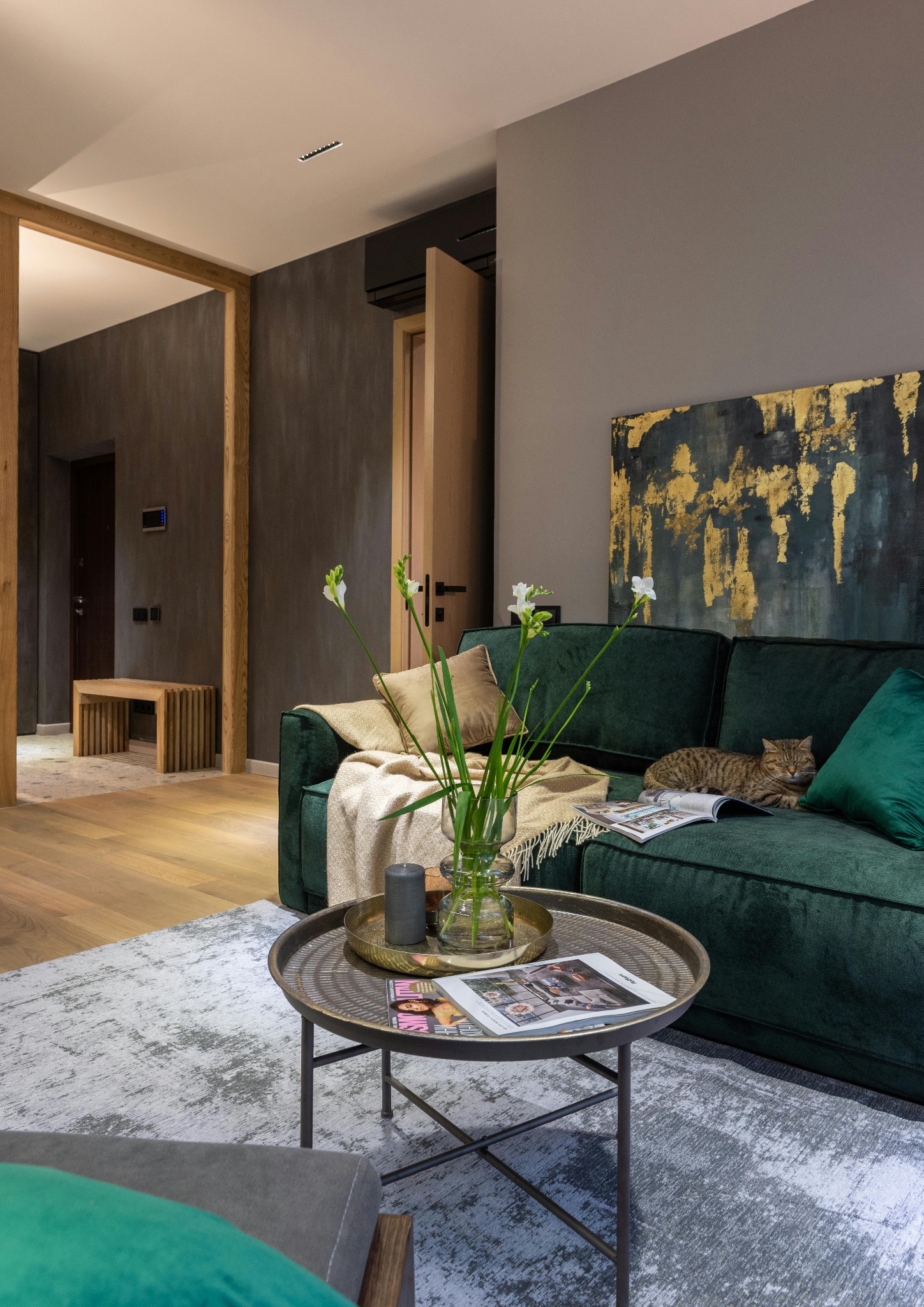 Stylowy salon z zieloną sofą, na której leży kot, stolik kawowy z wazonem i magazynem, oraz abstrakcyjny obraz na ścianie. Drewniane elementy dodają ciepła wnętrzu.
