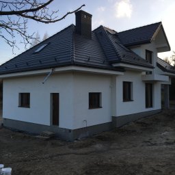 Remont Yaroslav Mazur - Nowo wybudowany, otynkowany dom jednorodzinny z ciemną dachówką, widoczne rury spustowe, teren wokół domu jeszcze niezagospodarowany, z przodu widoczne puszki z farbą.