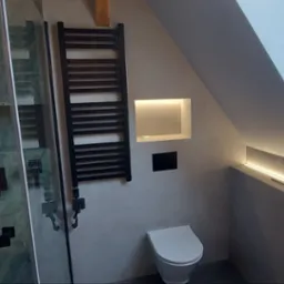 Nowoczesna łazienka na poddaszu z kabiną prysznicową wykończoną płytkami imitującymi marmur, grzejnikiem drabinkowym w kolorze czarnym, podświetlaną wnęką w ścianie i oknem dachowym.