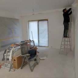 Remont Yaroslav Mazur - Pracownik na drabinie zabezpiecza folią malarską ścianę w jasnym, przygotowywanym do malowania pomieszczeniu; widoczne zakryte okno balkonowe i meble.