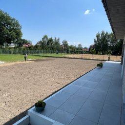 Remont Yaroslav Mazur - Przygotowanie terenu pod trawnik: wyrównana ziemia, widoczna osoba pracująca w oddali, obok ułożony chodnik z szarych płyt, doniczki z roślinami, błękitne niebo