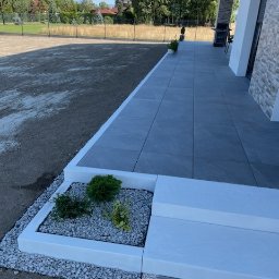 Remont Yaroslav Mazur - Nowoczesny taras z szarych płyt, białe obrzeża, rabata z krzewami i białym żwirem, widok na trawnik i dom.