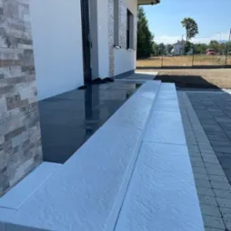 Nowoczesne wejście do domu z szarymi płytami i białymi stopniami, widok na świeżo ułożoną kostkę brukową i trawnik w tle.