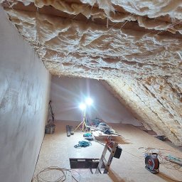 Remont Yaroslav Mazur - Izolacja poddasza wełną mineralną, widoczne narzędzia i drabina, oświetlenie halogenowe. Ściana boczna otynkowana, podłoga z płyt OSB.