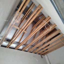 Remont Yaroslav Mazur - Konstrukcja drewniana poddasza z izolacją, widok z dołu w Bielsko-Białej. Elementy metalowe na suficie, przewody elektryczne. Prace wykończeniowe w trakcie.