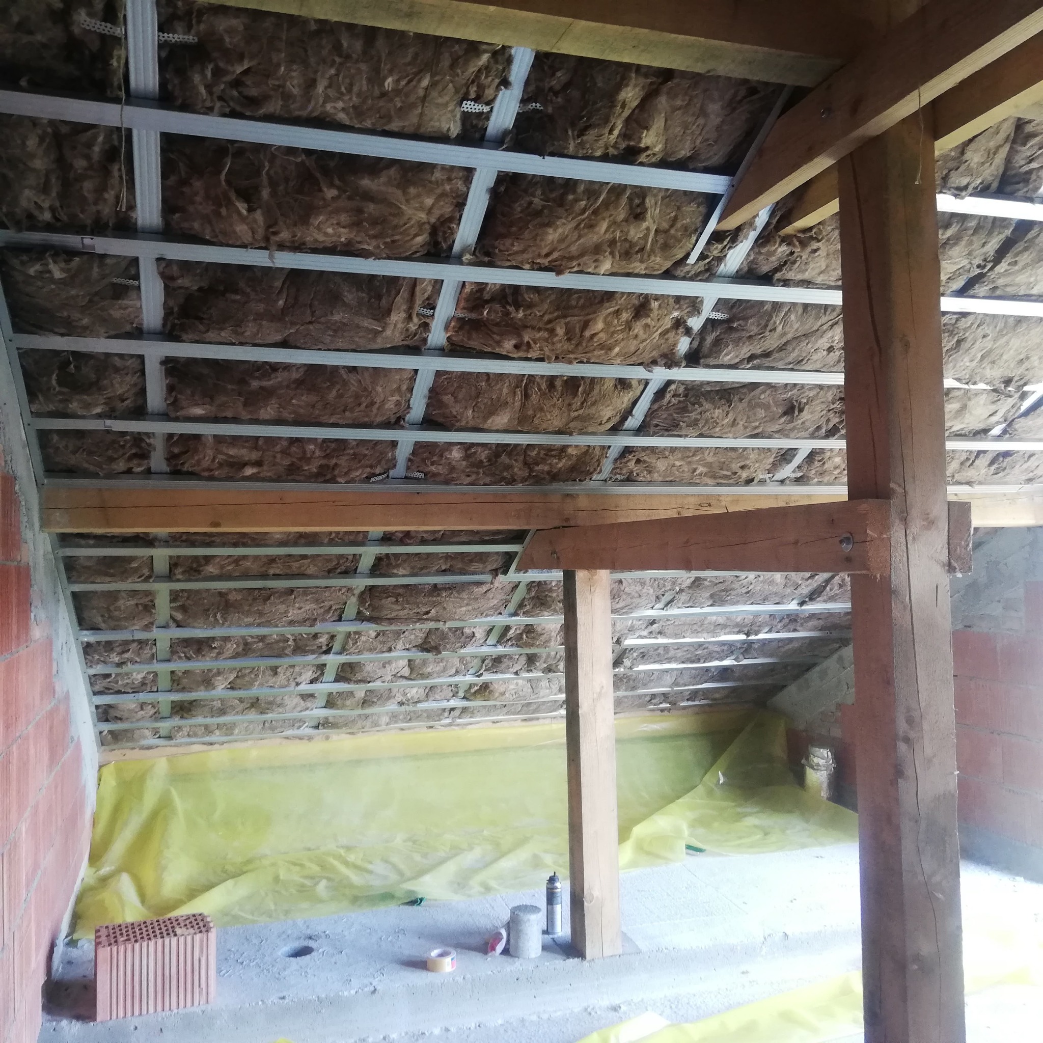 Izolacja poddasza wełną mineralną, widoczne profile metalowe, drewniane belki konstrukcyjne i folia ochronna na podłodze.