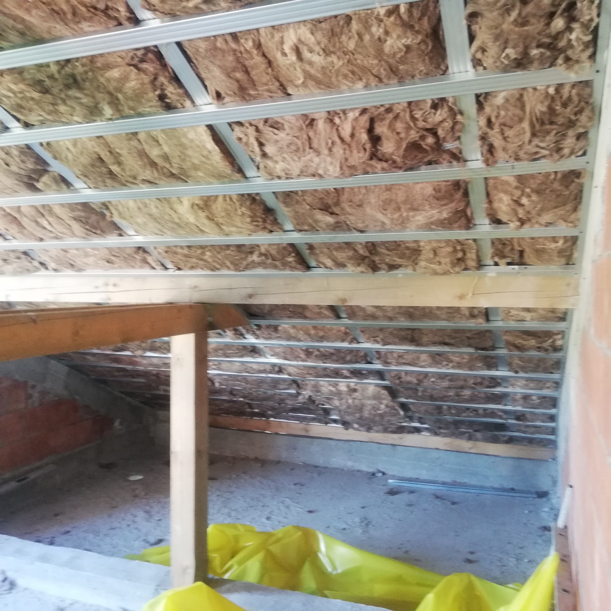 Izolacja poddasza wełną mineralną w metalowym stelażu, widoczne elementy konstrukcji drewnianej i folia ochronna na podłodze.