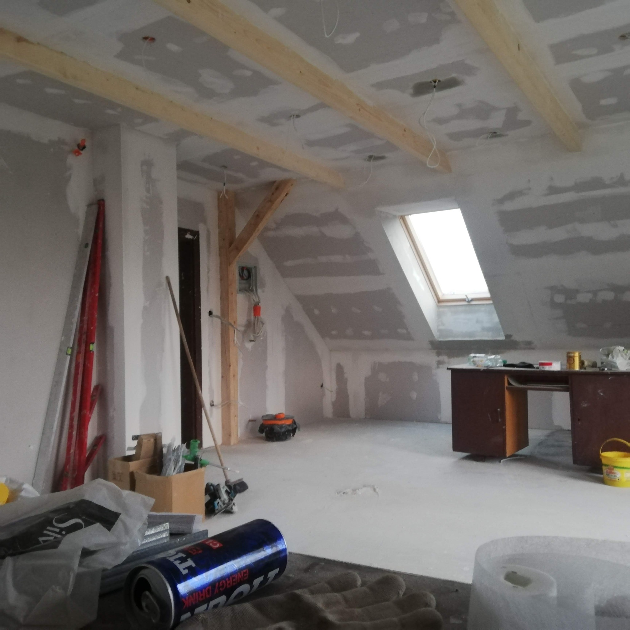 Poddasze w trakcie prac wykończeniowych, widoczne drewniane belki, okno dachowe, ściany pokryte płytami gipsowo-kartonowymi, narzędzia i puszka napoju energetycznego na pierwszym planie.