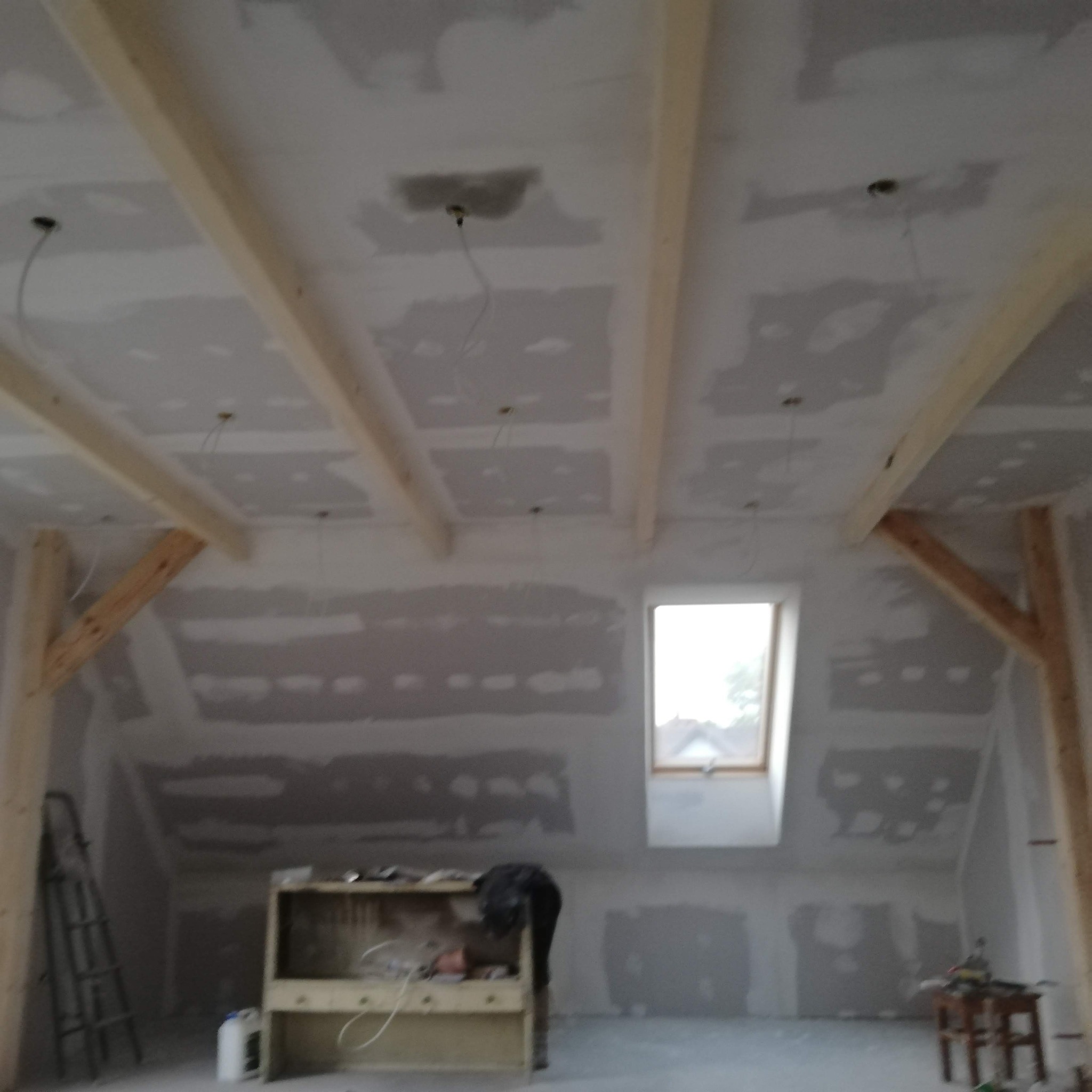 Poddasze w trakcie wykańczania: widoczne drewniane belki stropowe, płyty gipsowo-kartonowe na ścianach i suficie, okno dachowe, drabina oparta o ścianę oraz prowizoryczny warsztat z narzędziami.