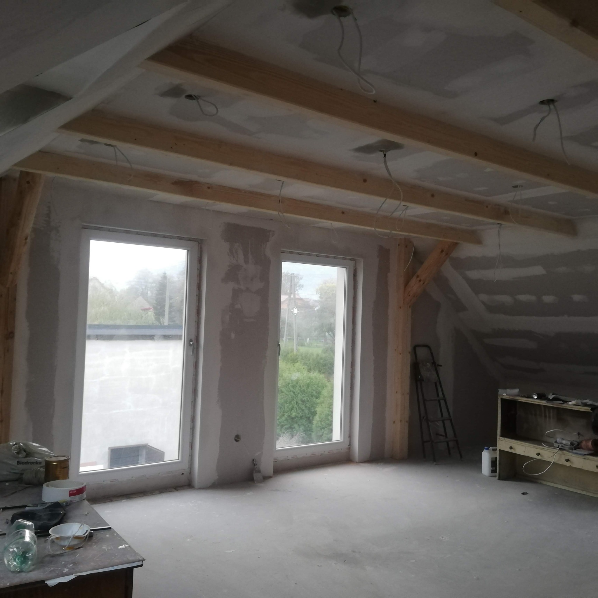 Poddasze w trakcie adaptacji: surowe ściany z gładzią, drewniana konstrukcja stropu z widocznymi kablami, okna PCV z widokiem na zewnątrz, drabina oparta o ścianę, narzędzia i materiały budowlane...