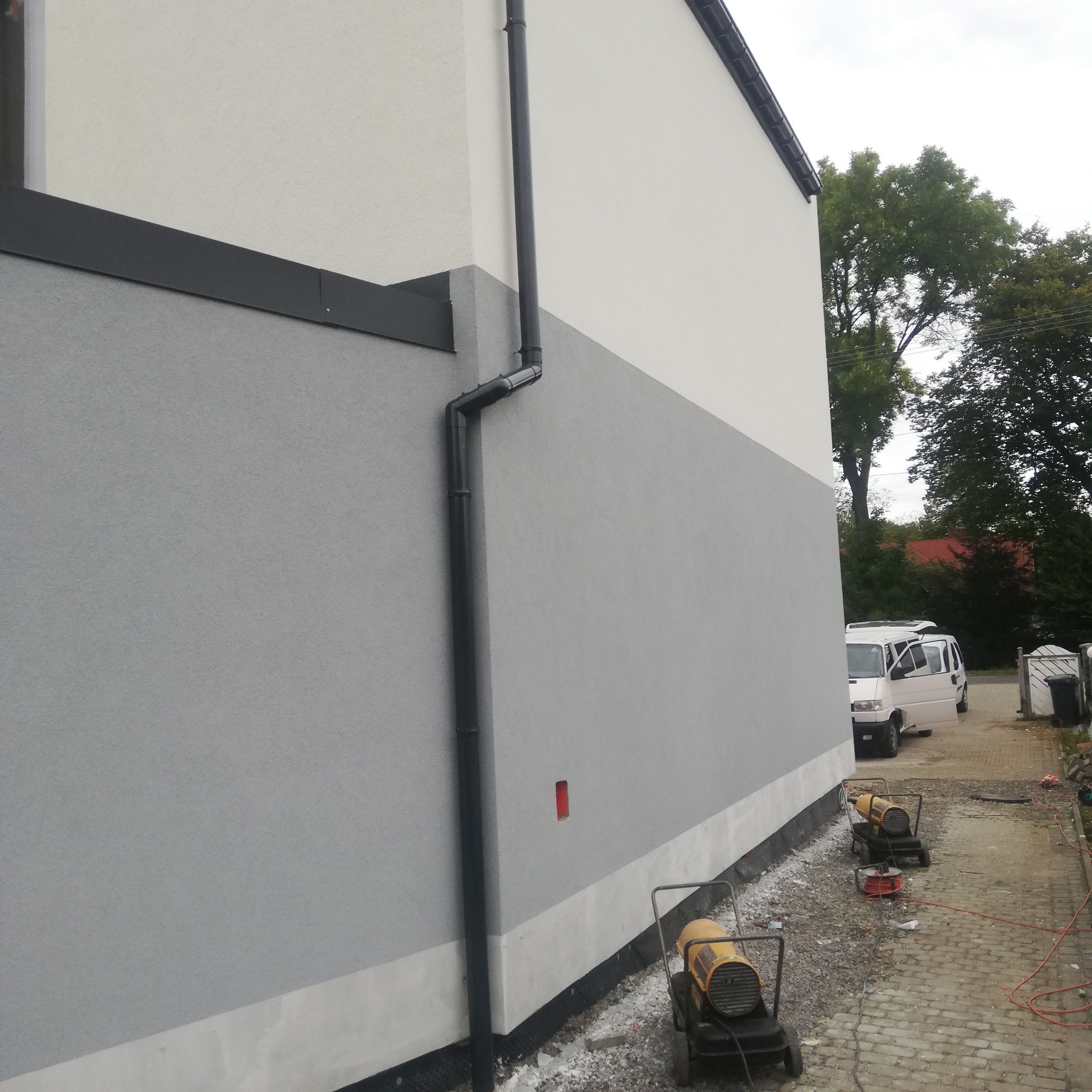 Świeżo otynkowana ściana budynku w Bielsku-Białej, z widocznymi dwoma nagrzewnicami budowlanymi i fragmentem chodnika z kostki brukowej. Na ścianie zamontowana rynna spustowa.