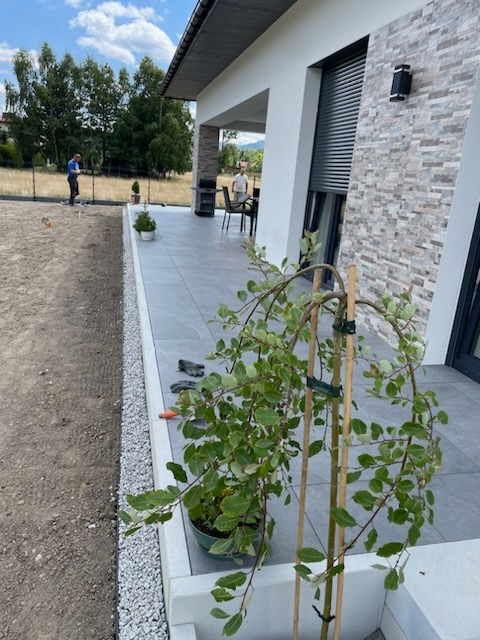 Nowoczesny taras z szarych płyt, obrzeże z białego grysu, trawnik w trakcie zakładania, widoczny człowiek z niwelatorem w tle.