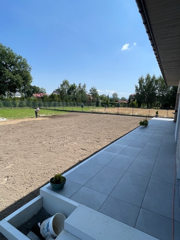 Przygotowanie terenu pod trawnik: wyrównana ziemia, widoczna osoba pracująca w oddali, obok ułożony chodnik z szarych płyt, doniczki z roślinami, błękitne niebo