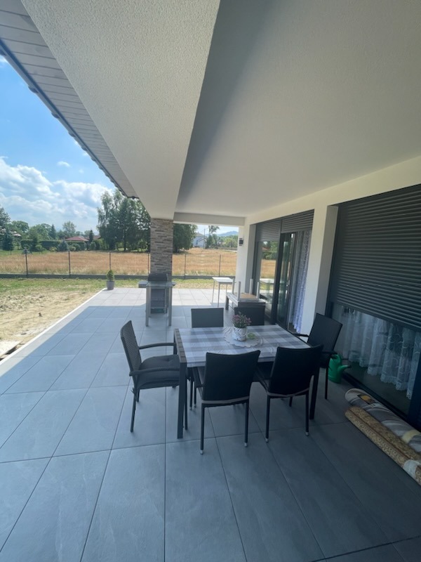 Zadaszony taras z szarych płytek, stół z krzesłami podparty kolumną z kamienia dekoracyjnego, widok na ogród z siatką.