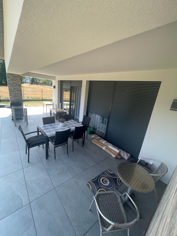 Zadaszony taras z szarymi płytkami, meblami ogrodowymi i zwiniętymi dywanami przy ścianie z żaluzjami.