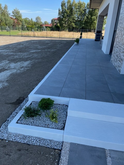 Nowoczesny taras z szarych płyt, białe obrzeża, rabata z krzewami i białym żwirem, widok na trawnik i dom.