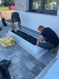 Dwóch mężczyzn w trakcie układania czarnej folii w białej, prostokątnej donicy na kostce brukowej przed budynkiem z białym murem. W tle widoczny czerwony samochód dostawczy i myjka ciśnieniowa.