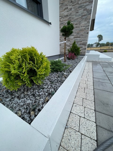 Kaskadowa rabata z roślinami iglastymi i ozdobnymi kamykami, obrzeże z białego betonu, kostka brukowa w dwóch odcieniach szarości, nowoczesna architektura domu w tle.