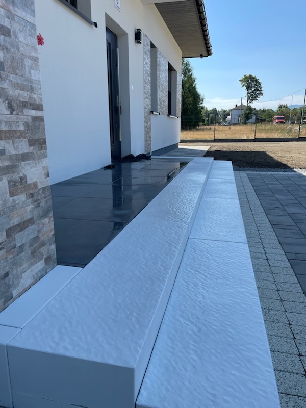 Nowoczesne wejście do domu z szarymi płytami i białymi stopniami, widok na świeżo ułożoną kostkę brukową i trawnik w tle.