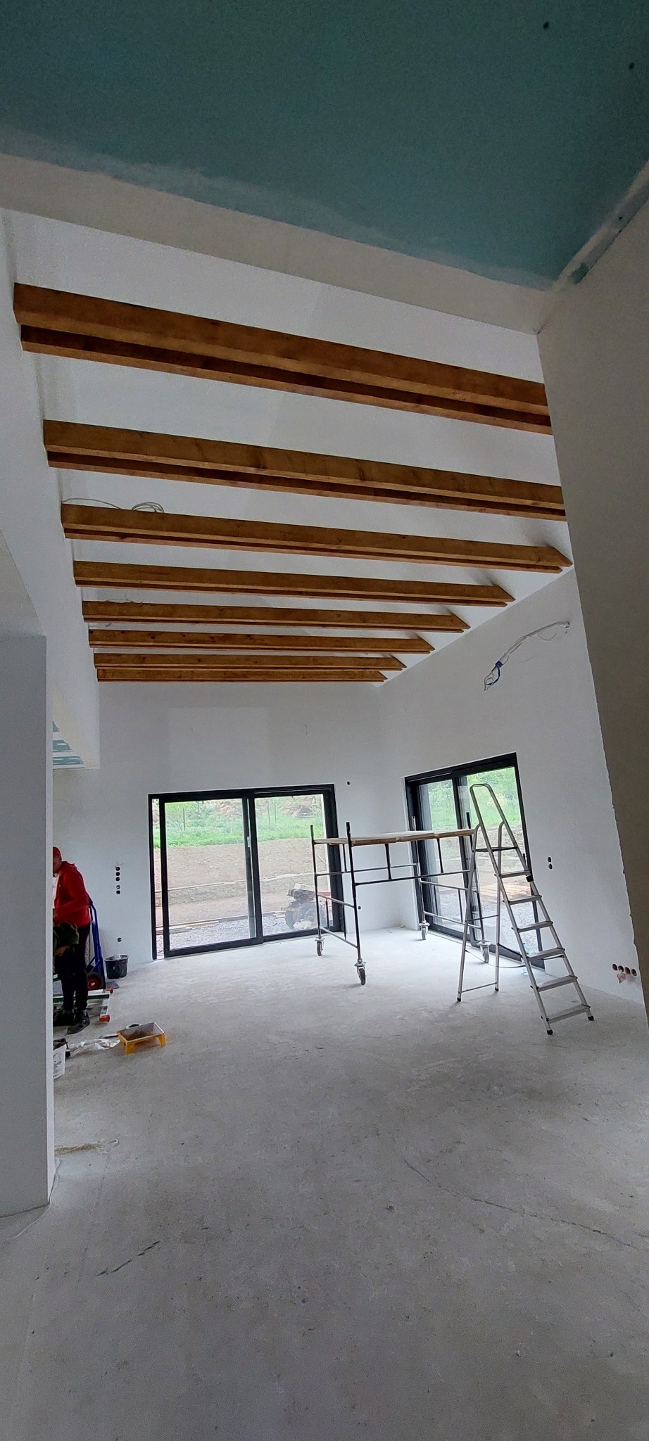 Wnętrze w trakcie wykończenia: surowe ściany, drewniane belki stropowe, okna przesuwne, rusztowanie i drabina. Widoczna osoba w czerwonej bluzie.