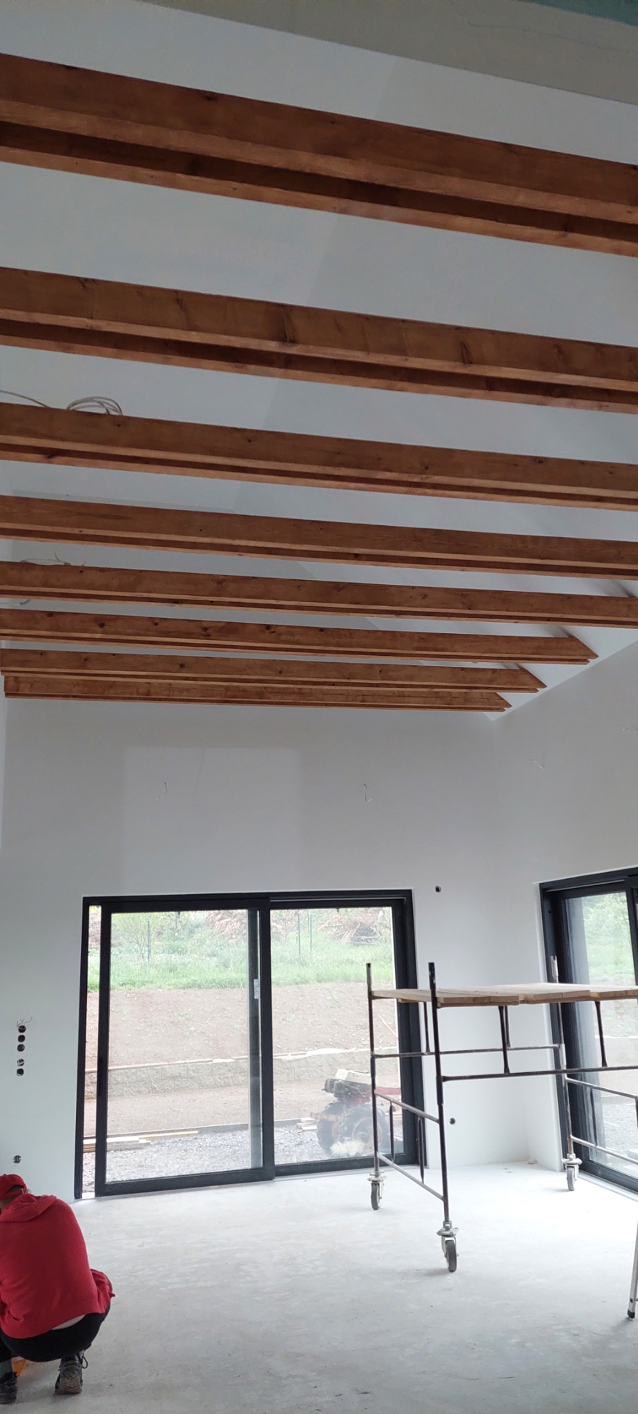 Wnętrze w trakcie wykończenia: białe ściany, drewniane belki stropowe, okna przesuwne z czarnymi ramami, rusztowanie na kółkach i osoba w czerwonej bluzie w rogu.