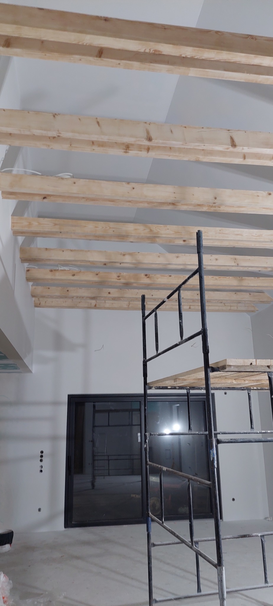 Montaż drewnianej konstrukcji sufitu podwieszanego z widocznym rusztowaniem i oknem w tle. Prace wykończeniowe w nowym budynku.