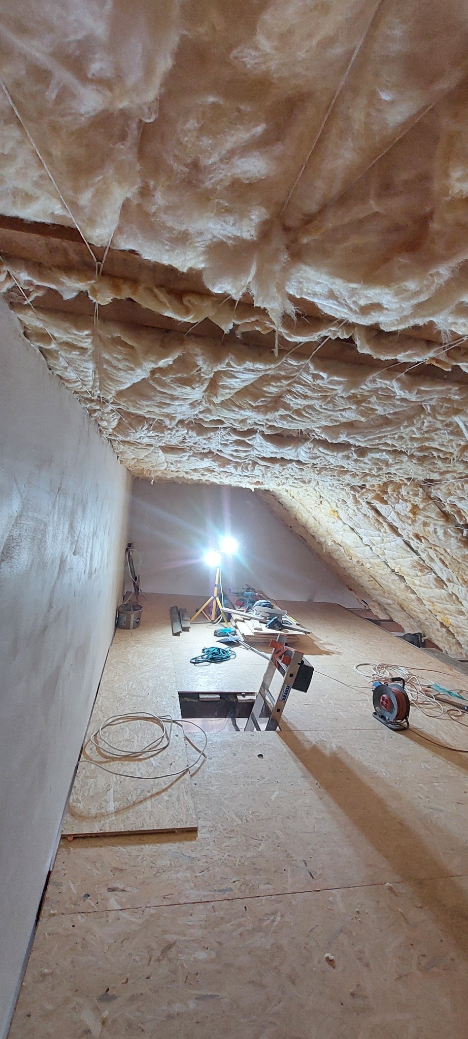 Izolacja poddasza wełną mineralną, widoczne narzędzia i drabina, oświetlenie halogenowe. Ściana boczna otynkowana, podłoga z płyt OSB.