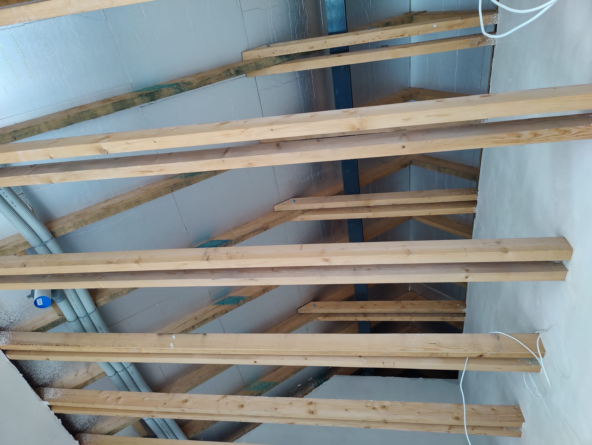 Skośny kadr na drewnianą konstrukcję poddasza z izolacją folią aluminiową i fragmentami instalacji. Widoczne elementy więźby dachowej i docieplenia.