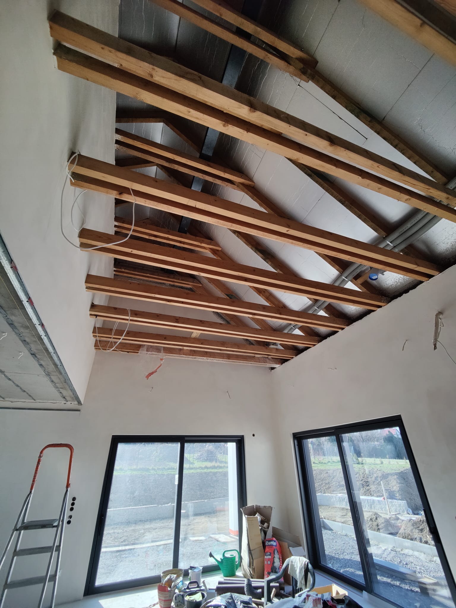 Wnętrze w trakcie wykończenia: drewniana konstrukcja dachu z izolacją, surowe ściany, duże okna z widokiem na zewnątrz, drabina oparta o ścianę, narzędzia i materiały budowlane.
