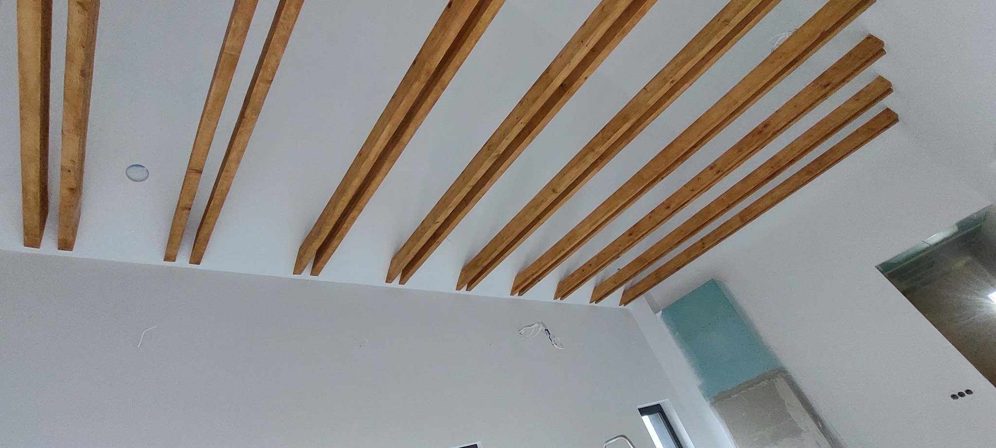 Drewniane belki dekoracyjne pod sufitem w jasnym wnętrzu. Widoczne fragmenty ścian i okna. Surowy, nowoczesny design.