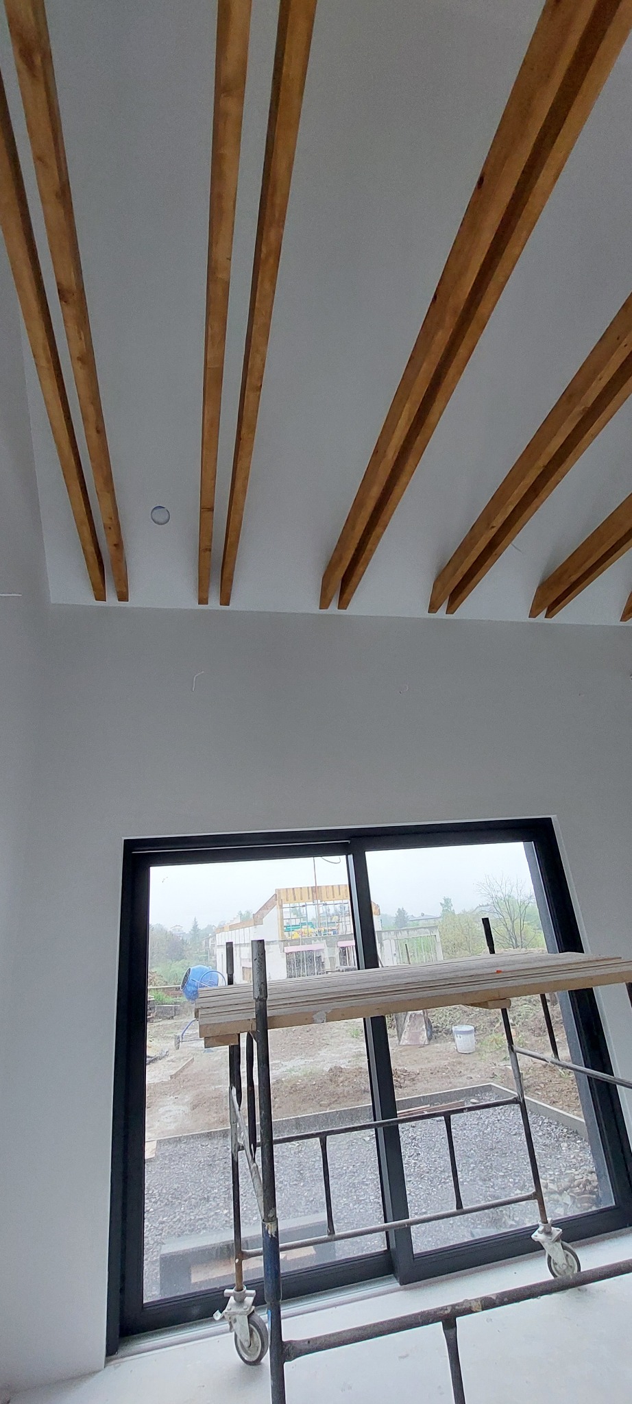 Drewniane belki stropowe w nowoczesnym wnętrzu, widok z okna na plac budowy. W tle rusztowanie i materiały budowlane.