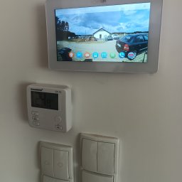 ARTELMET - Monitor wideodomofonu zamontowany na ścianie, wyświetlający obraz z kamery na zewnątrz, obok termostat i włączniki światła.