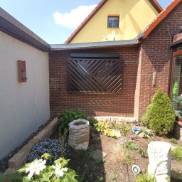 ARTELMET - Roleta zewnętrzna z dekoracyjnym wzorem na ceglanej ścianie budynku, widok na fragment ogrodu z roślinami i elementami dekoracyjnymi.