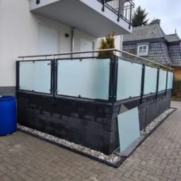 Nowoczesna balustrada ze szkła matowego i czarnych profili na tarasie przed domem z kostki brukowej, widoczny fragment niezamontowanej szyby.