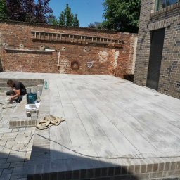 ARTELMET - Układanie jasnoszarej kostki brukowej na tarasie z ceglanym murem w tle, widoczny pracownik z narzędziami.