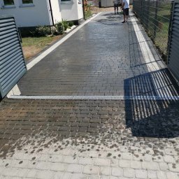 ARTELMET - Czyszczenie kostki brukowej na podjeździe przed domem; widoczne ślady wody i różnica w kolorze pomiędzy czystą a brudną powierzchnią, metalowy odpływ liniowy, częściowo otwarta brama wjazdowa...