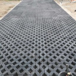 ARTELMET - Ułożona nawierzchnia z szarych, ażurowych płyt betonowych na podjeździe, widoczne narzędzia w wiadrze i piasek na brzegach.